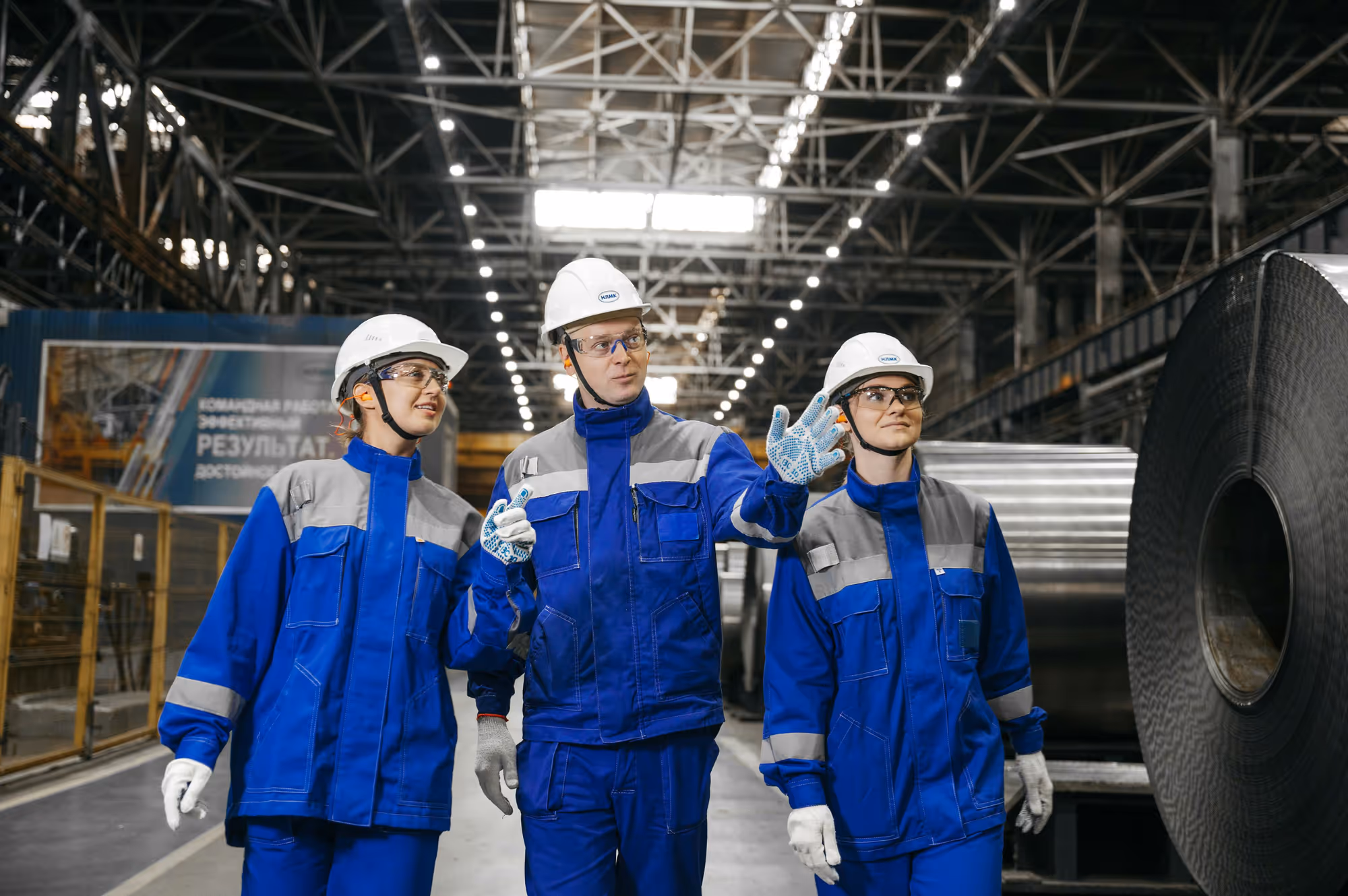 Steelworkers in protective equipment walking a mill floor beside a coil of strip steel.