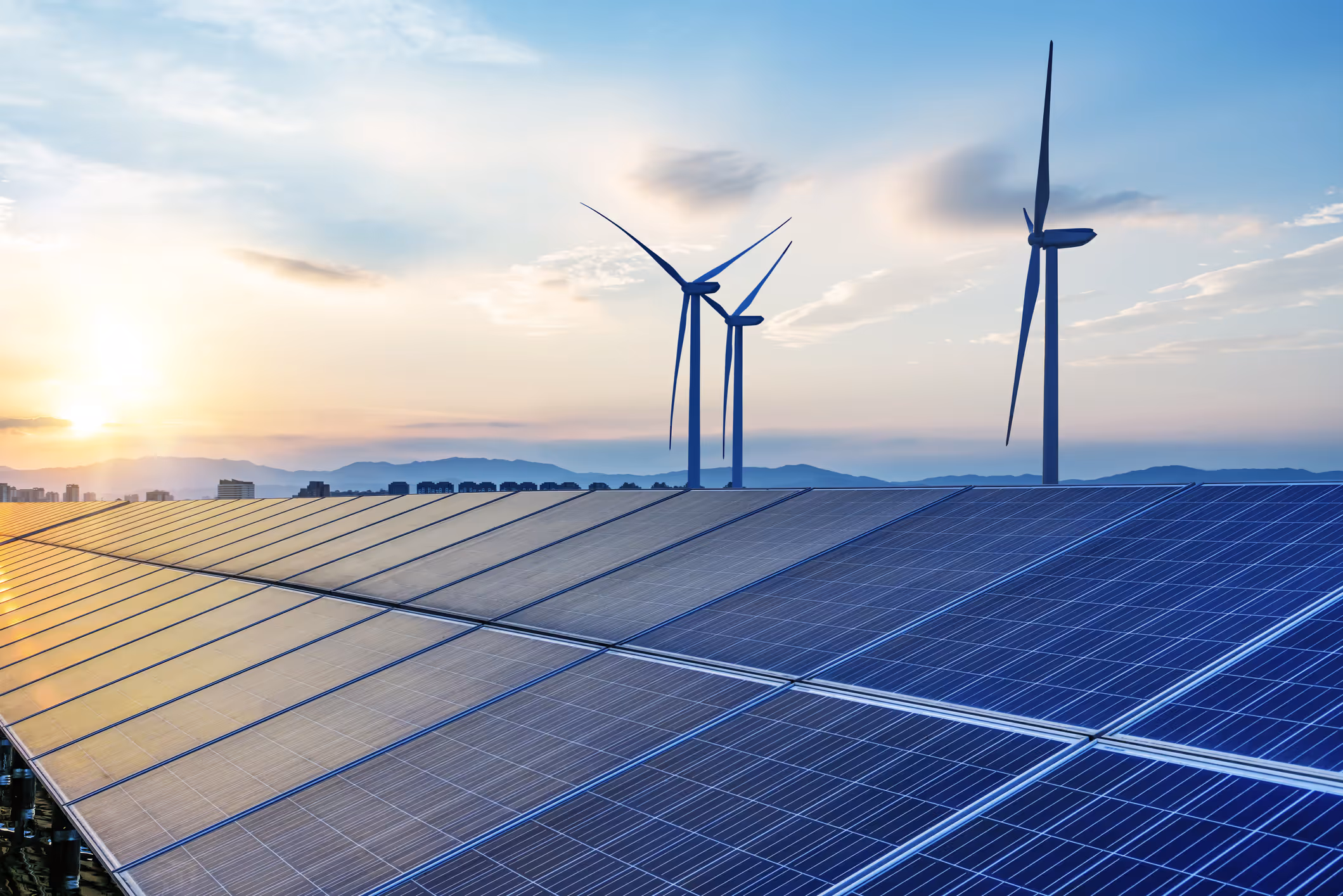 Solar panels in the foreground with wind turbines rising behind them at sunrise.